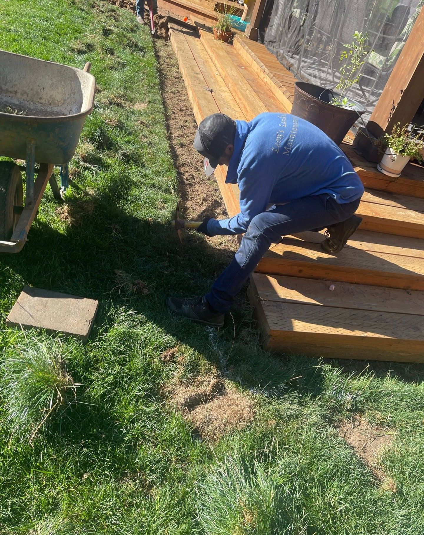Man kneeling, working on edge of wooden deck next to grass. Wheelbarrow nearby.