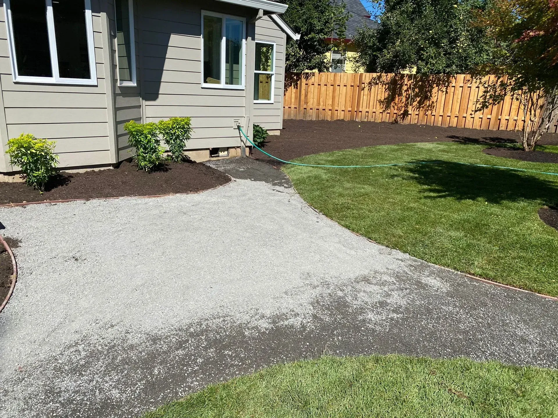 Gravel pathway leads to a light grey house with shrubs, bordered by grass and mulch. Brown fence in the background.