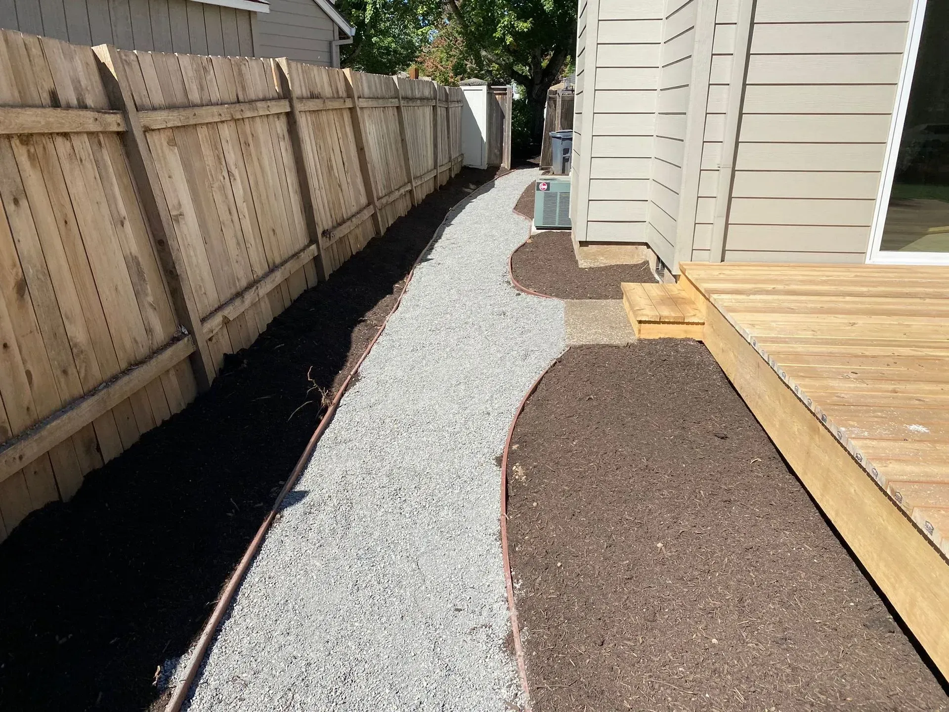 Gravel path alongside a wooden fence and a house with a wooden deck; dark mulch borders the path.