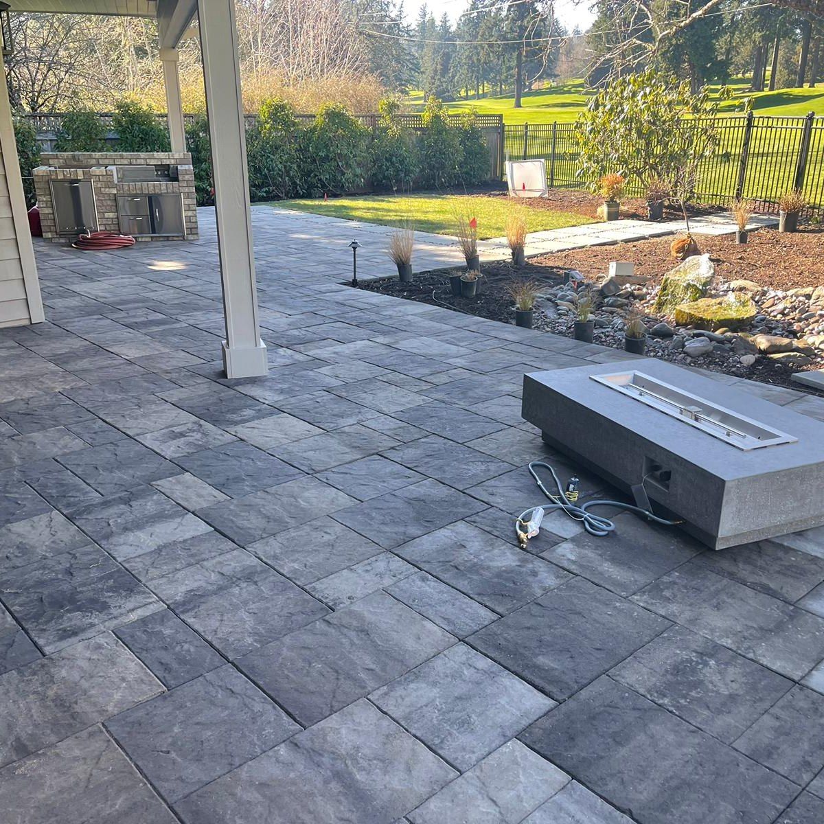 Patio paved with gray blocks, with a fire pit, overlooking a grassy yard with landscaping.