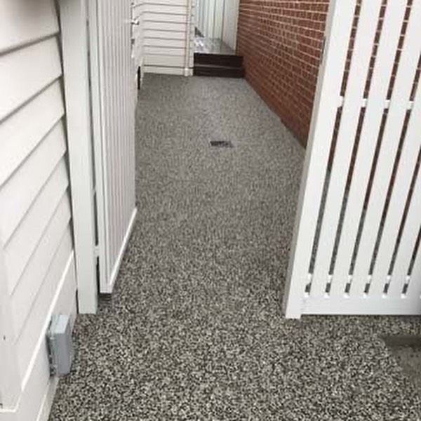 Narrow outdoor pathway with speckled gray flooring, between white slatted fences and a brick wall.