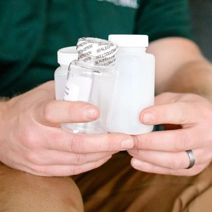 Hands holding three small plastic bottles. One is clear, the others white. All have white lids.