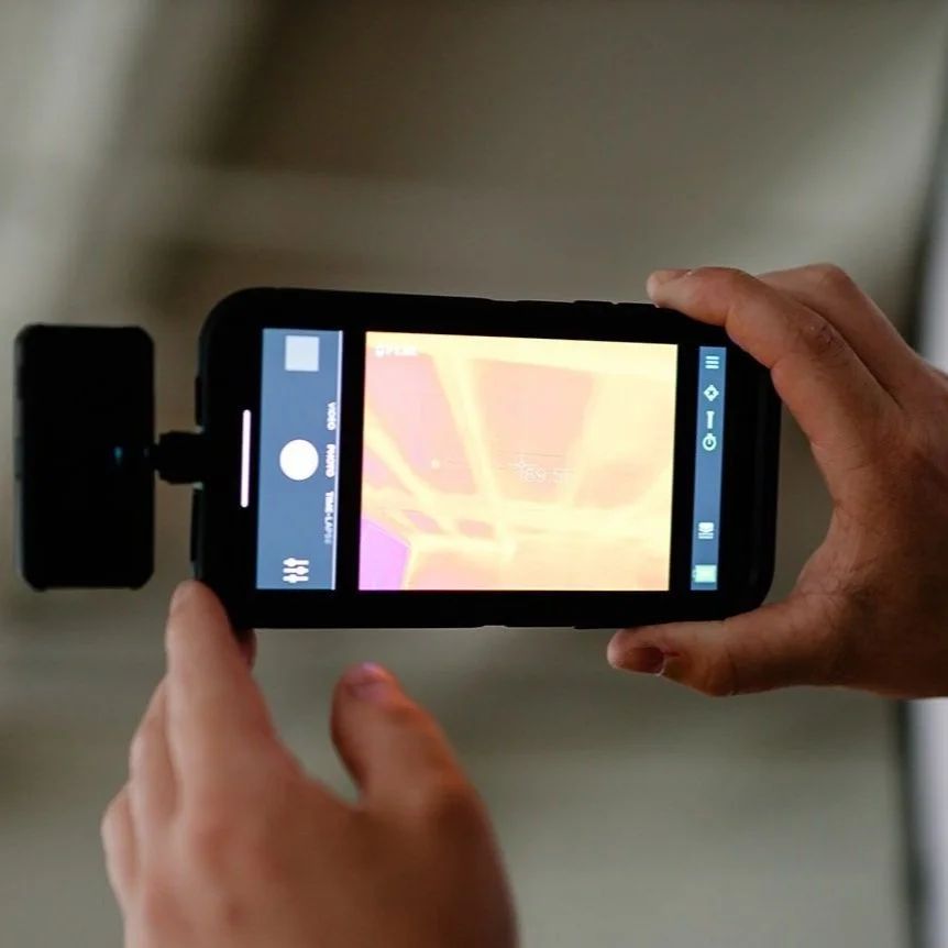Hands holding a smartphone with a thermal camera attached, displaying a heatmap of an object.