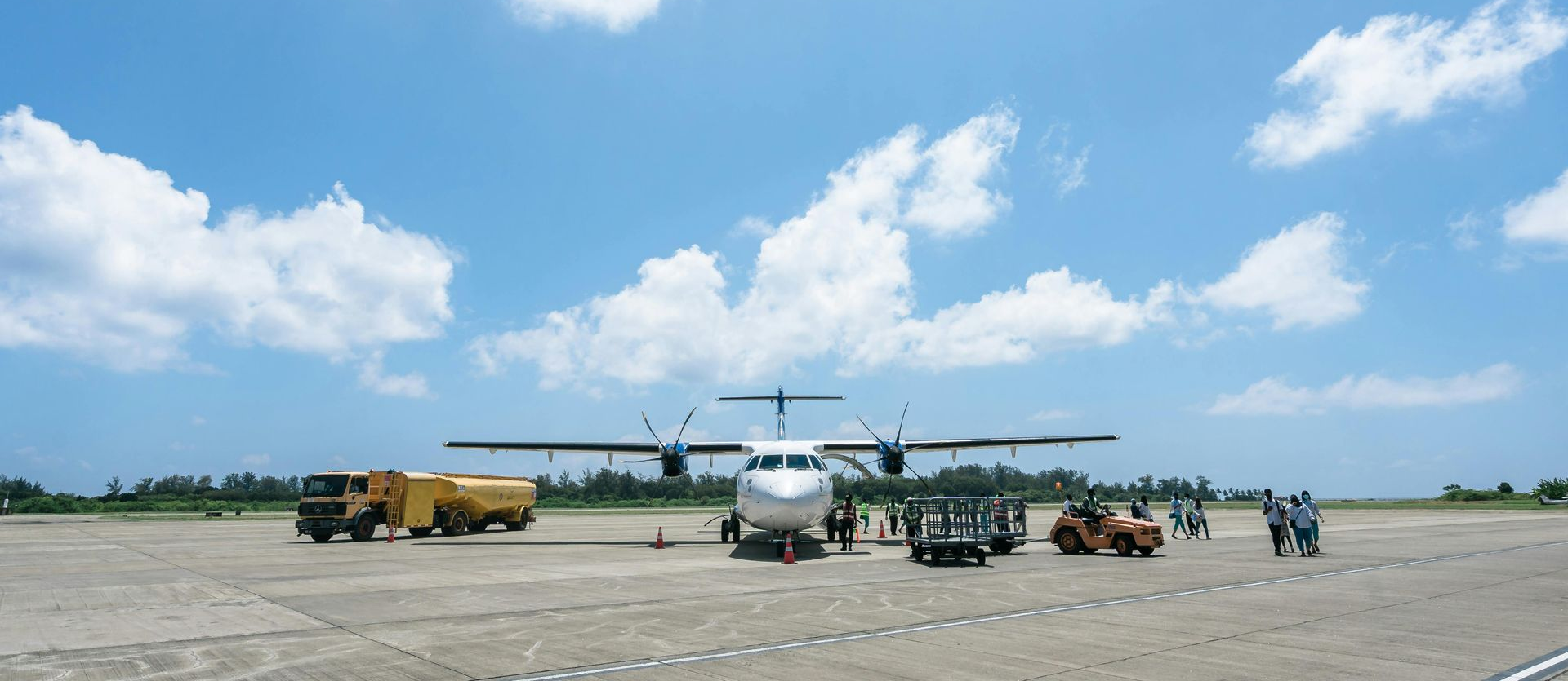 Airplane on a tarmac with service vehicles and people under a bright blue sky with clouds.