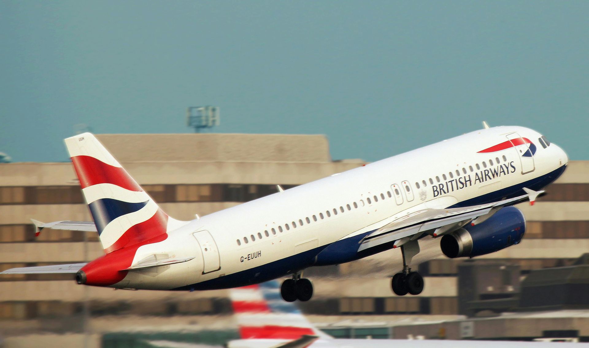 British Airways plane taking off, white and blue, with a Union Jack tail, near an airport building.