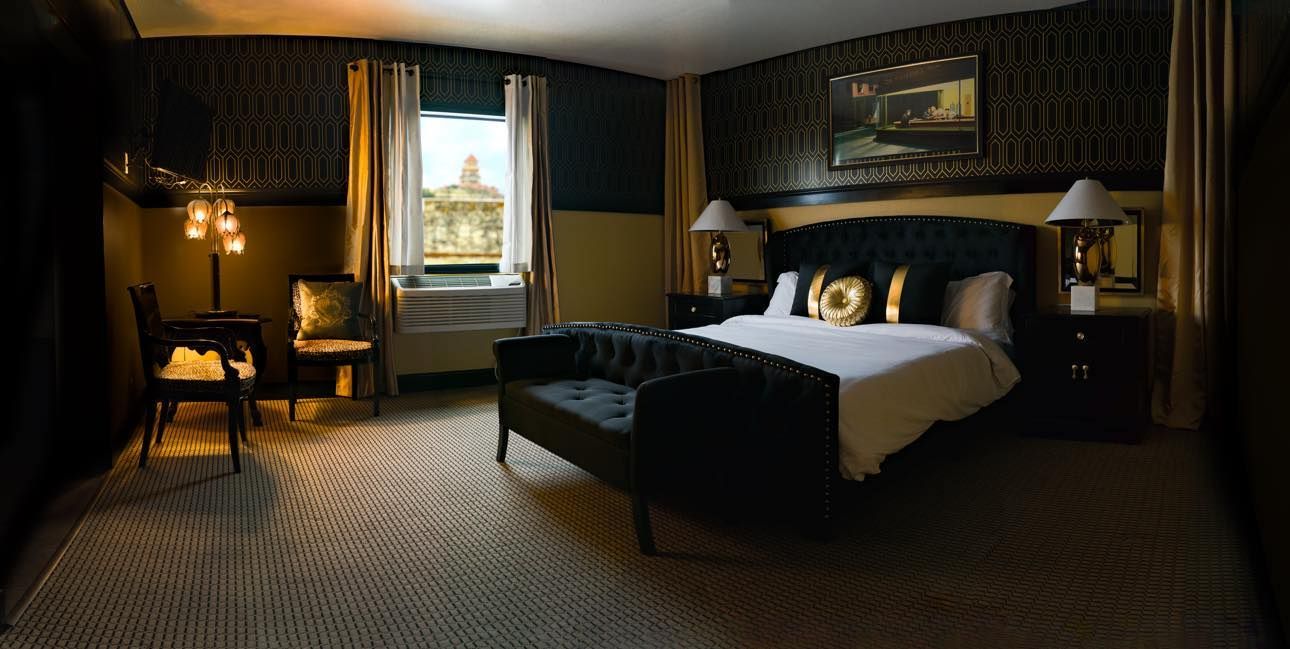 A hotel room with a King bed , chairs , lamps and a window.