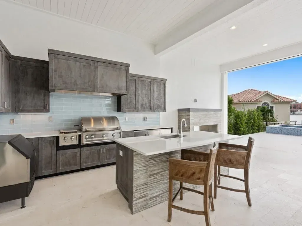 Modern outdoor kitchen with island, grill, wood cabinets, and open view to a backyard and house.