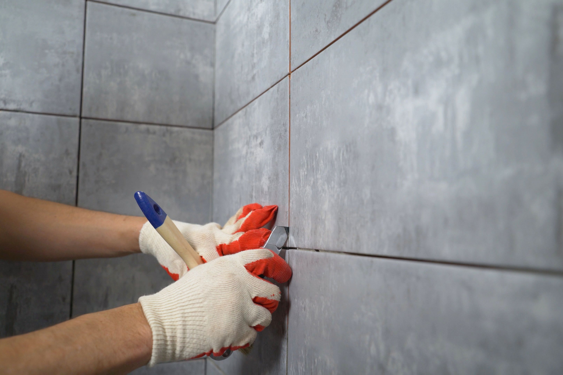 Hands wearing gloves apply grout to gray wall tiles with a blue-and-white tool.