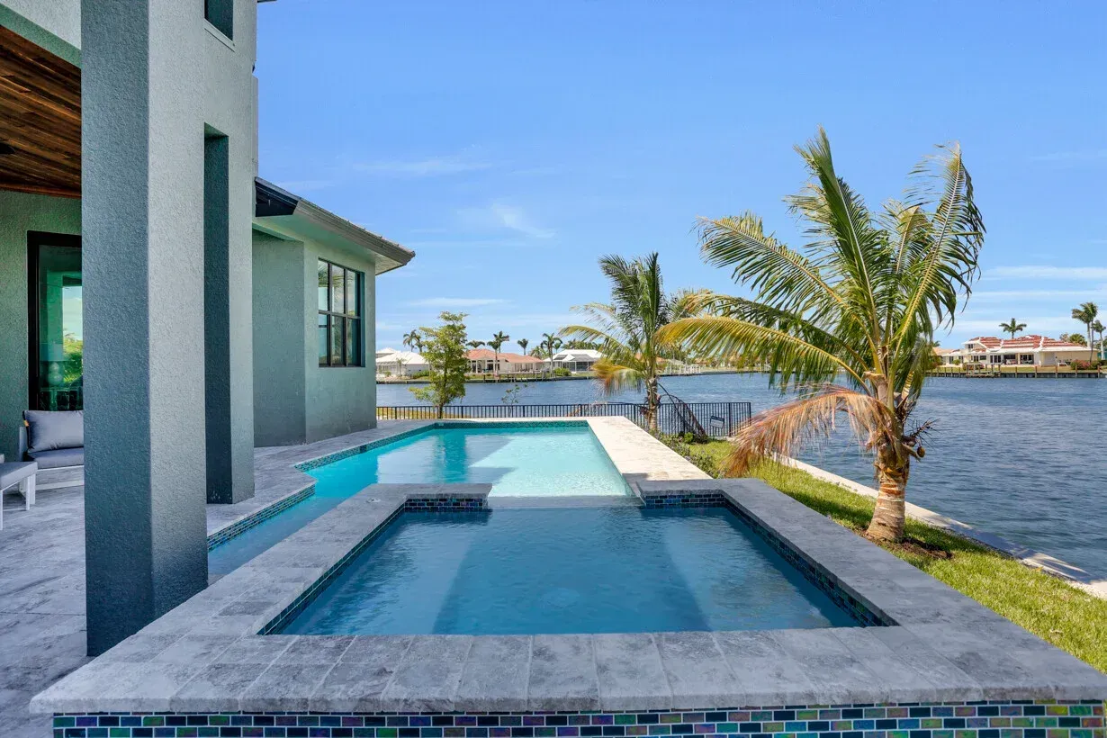 Modern waterfront patio with a small pool, palm trees, and a house beside a calm bay.