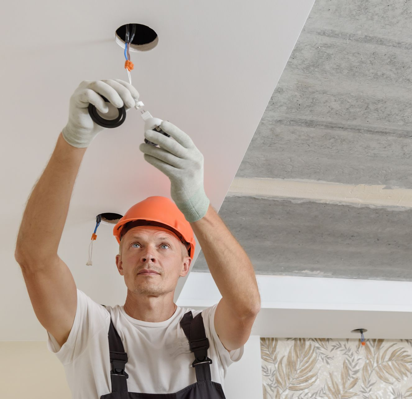 Worker installing a ceiling light fixture with tools and safety gear on a renovation site.