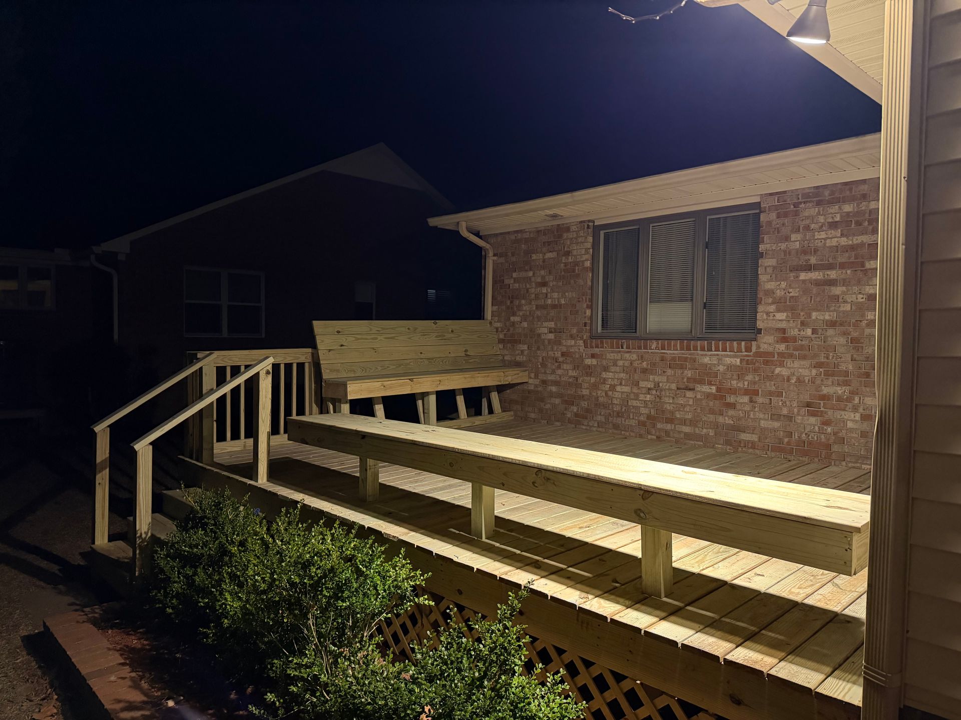Wooden deck with a built-in bench, next to a brick building, lit by outdoor lights at night.