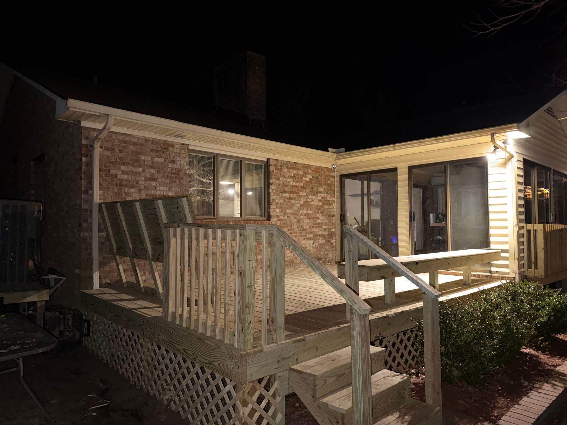 Wooden deck with steps, connected to a brick house with sliding glass doors, at night.