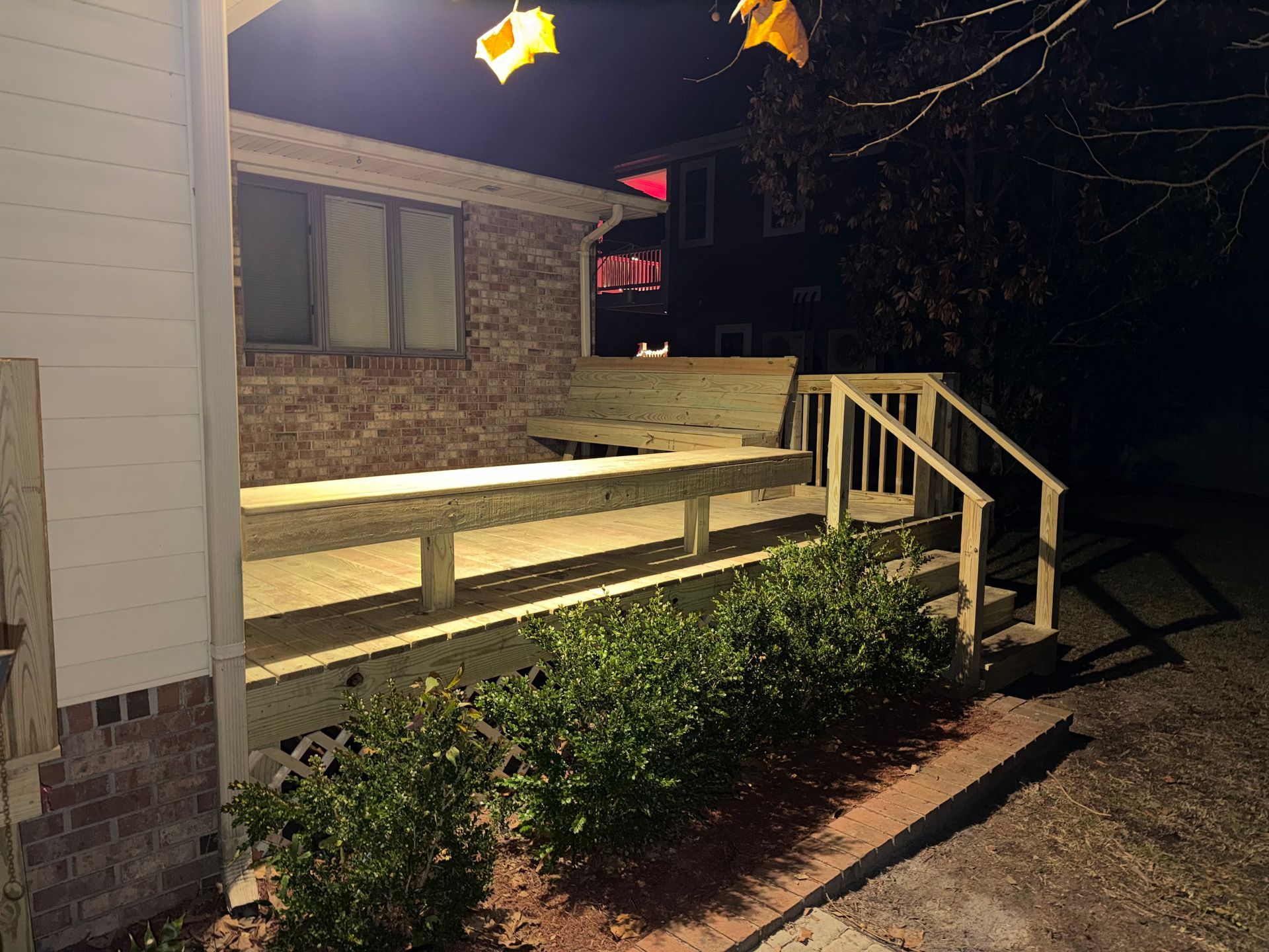 A wooden deck illuminated by a light at night, with bushes lining the edge, beside a brick and white-sided house.