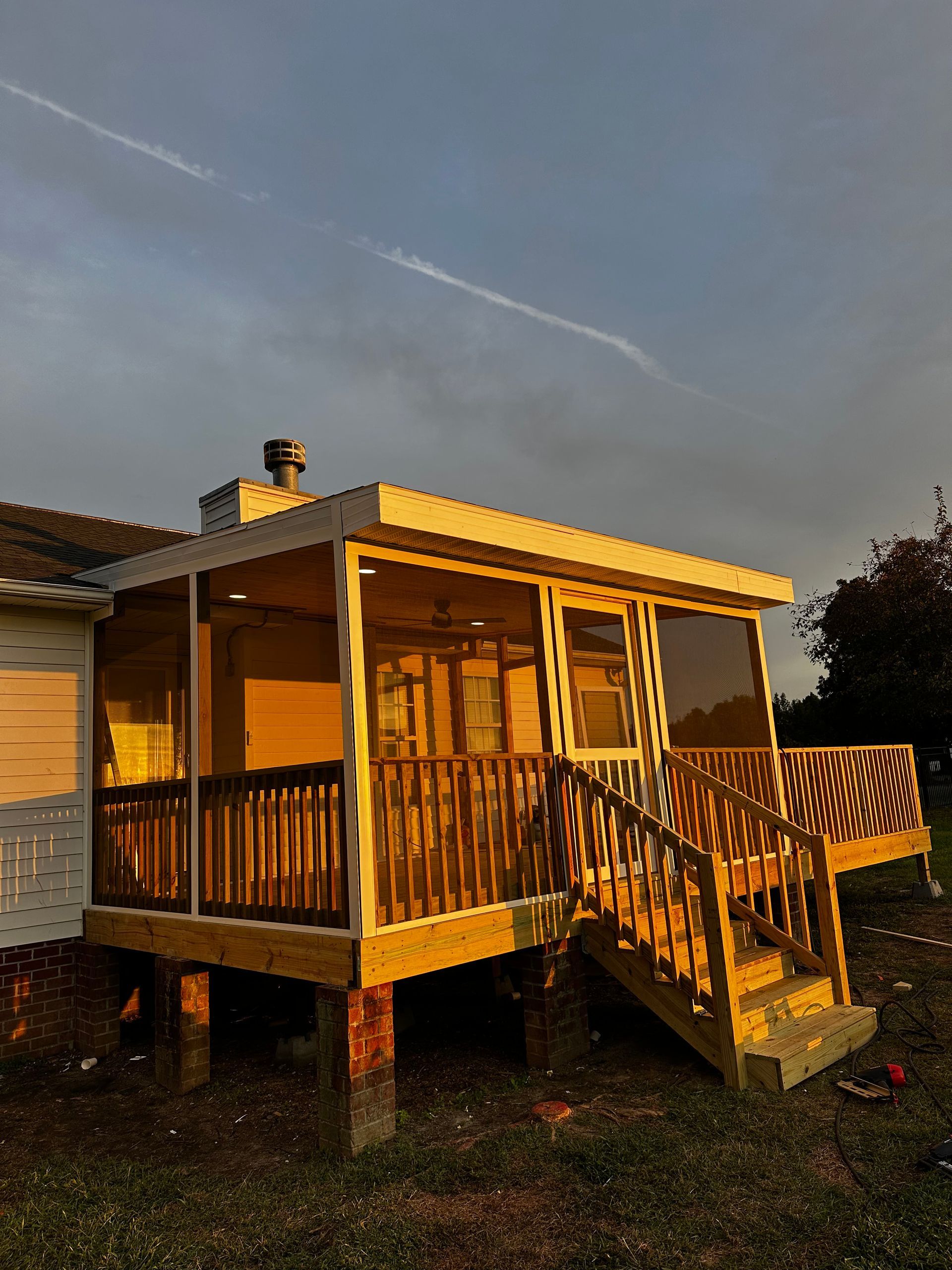 Wooden screened porch with stairs and deck, attached to a house, lit by golden sunlight.