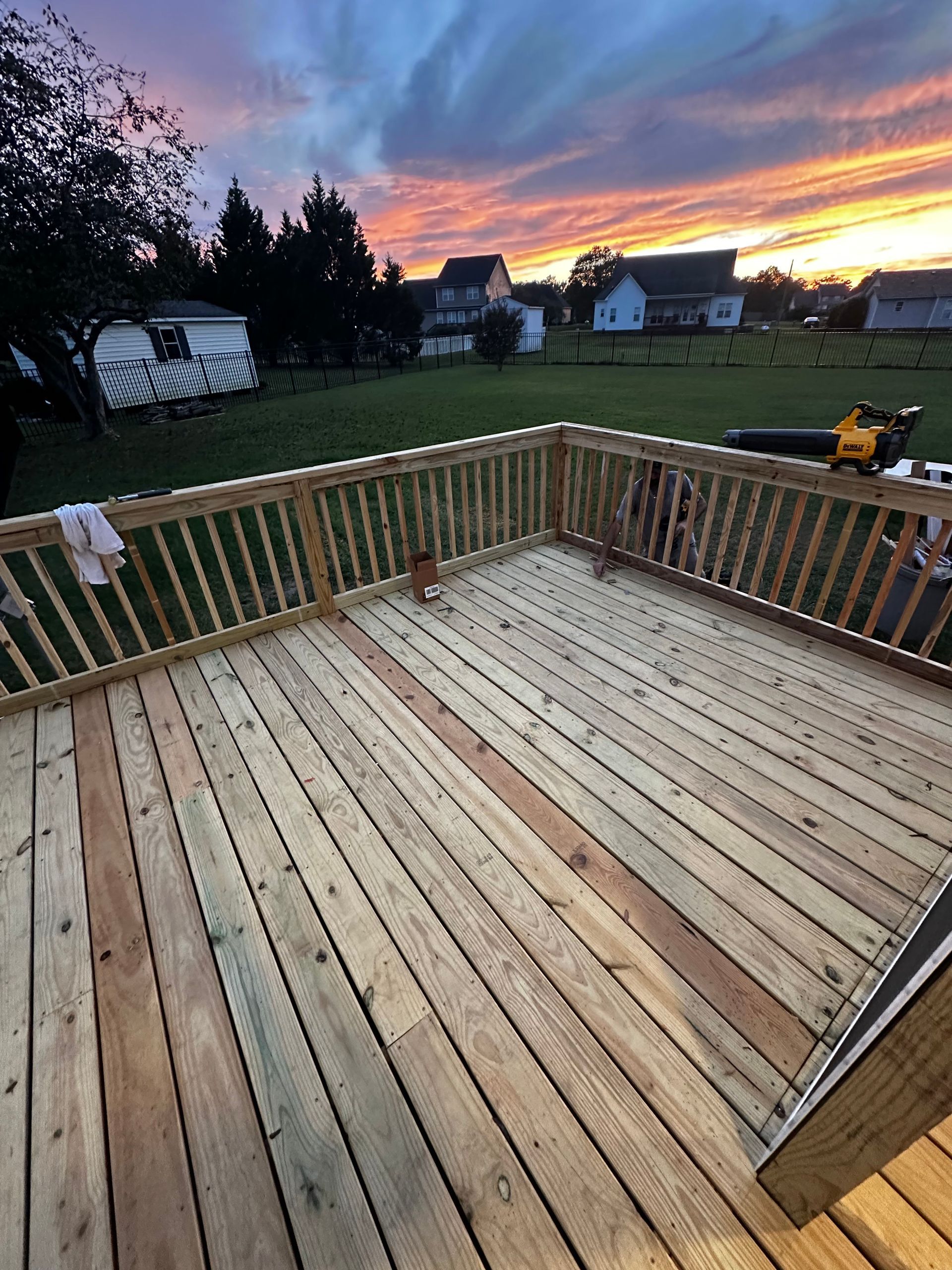 Wooden deck with railing, lawn, houses, and colorful sunset in background.