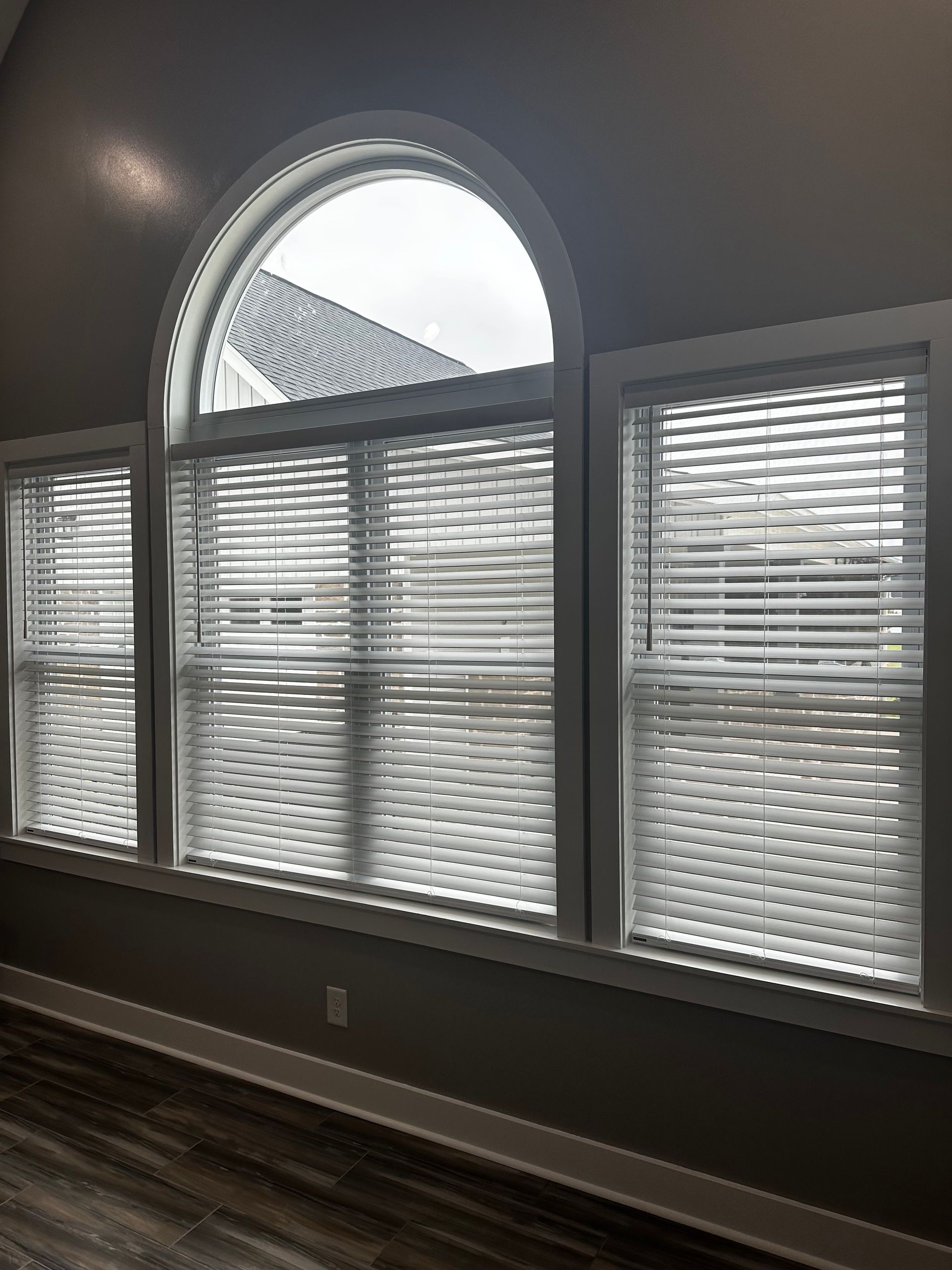 Three windows with white frames and blinds; one arched, overlooking a building, set in a gray room.