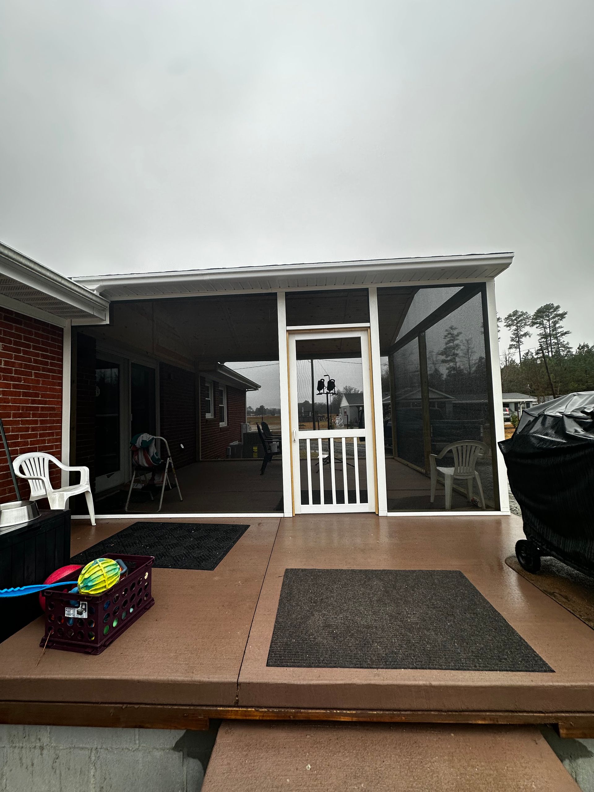 Screened-in porch attached to a brick house. Door and windows with black screening, brown deck, and gray sky.