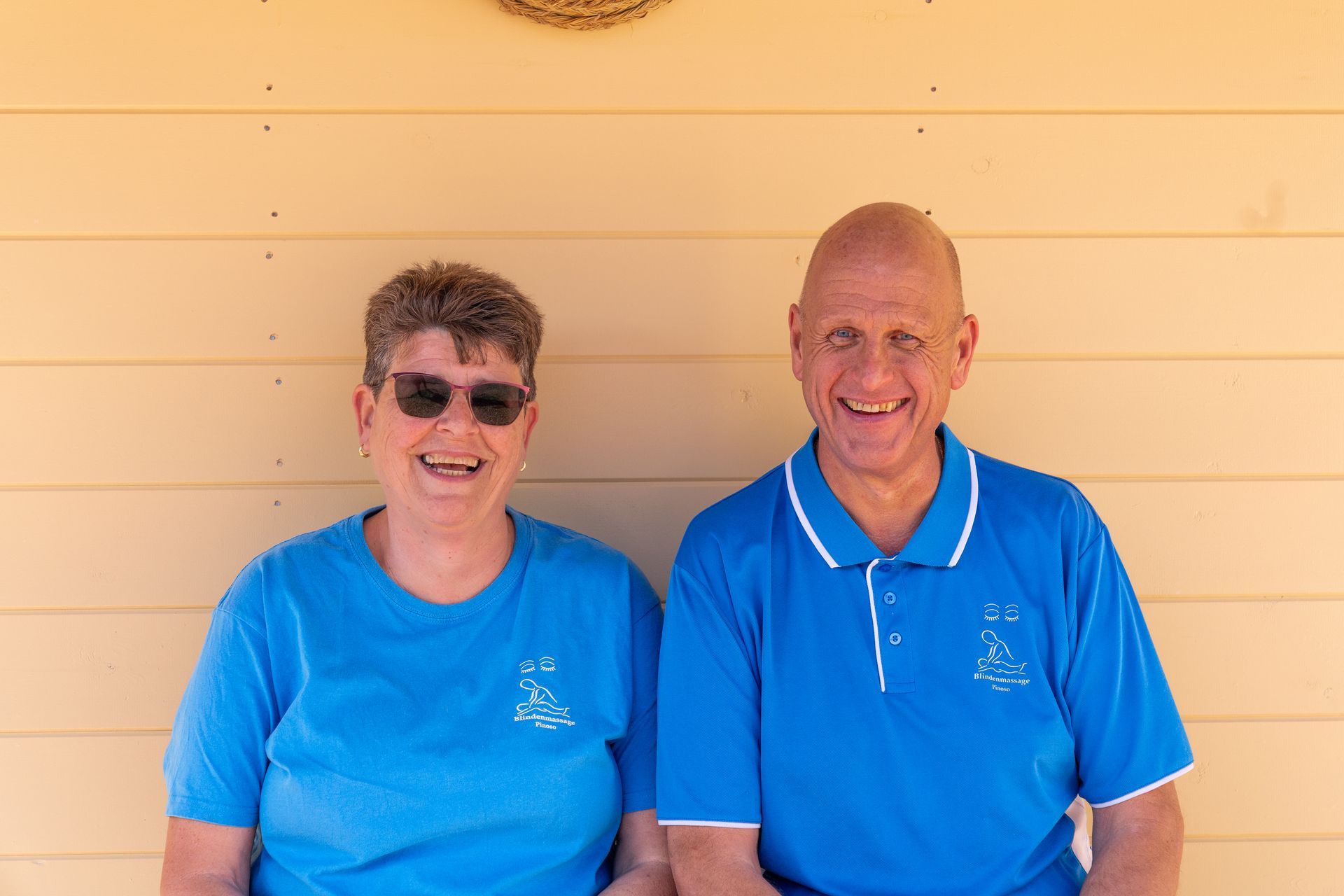 Twee lachende mensen in blauwe shirts staan ​​voor een beige muur.