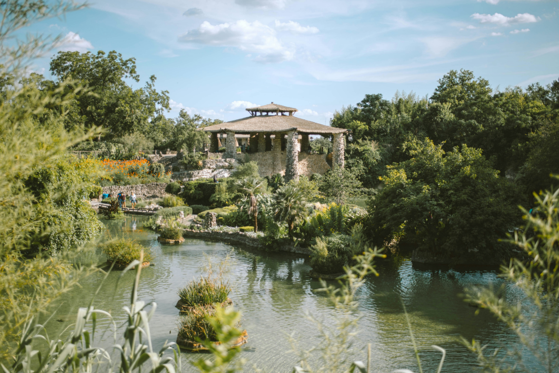 Lush garden pond with a gazebo, flowering plants, and dense green trees under a blue sky.