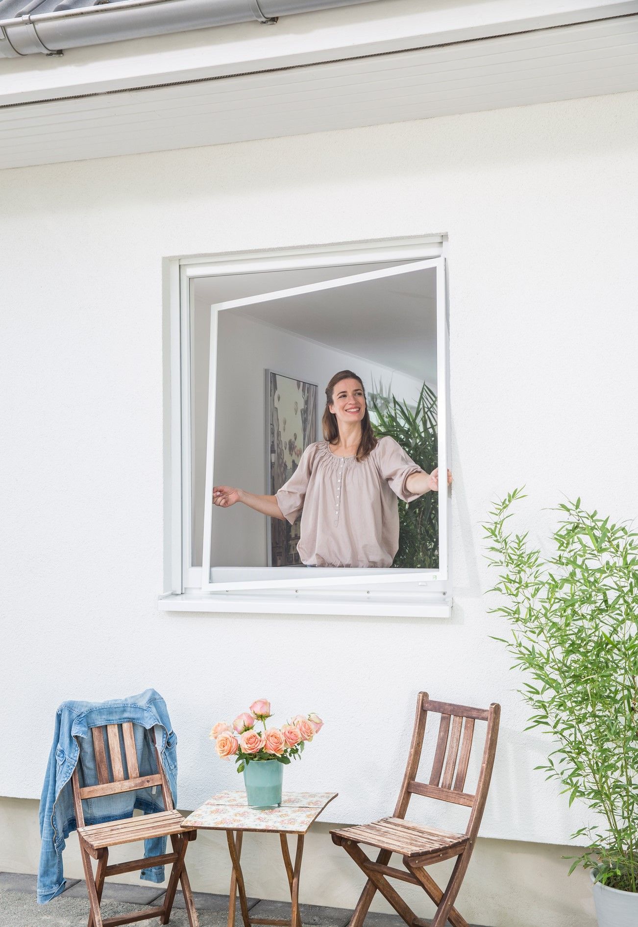 Femme à la fenêtre avec écran ouvert, souriante ; décor extérieur avec table, chaises, fleurs et veste.