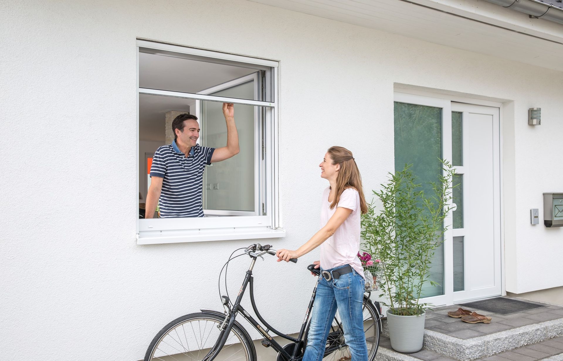 Un homme à la fenêtre ajuste l'écran, une femme avec un vélo sourit à l'extérieur d'une maison blanche.