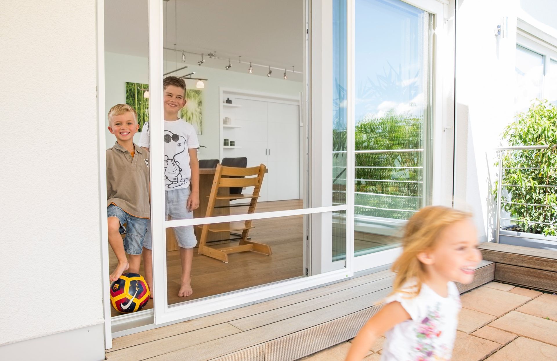 Deux garçons et une fille devant une porte coulissante en verre avec moustiquaire. Un enfant court et les garçons sourient.