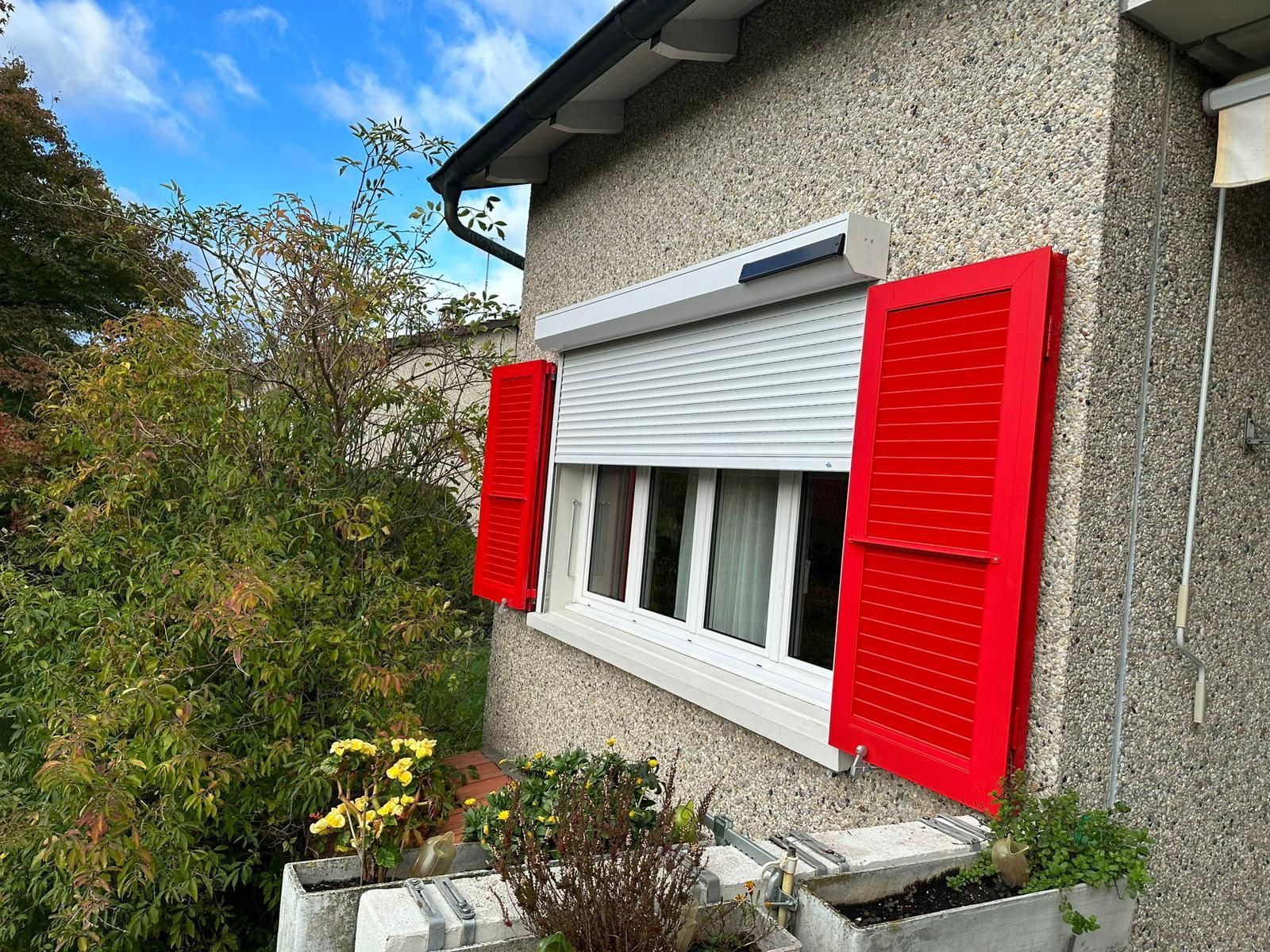 Des volets rouges encadrent une fenêtre avec un volet roulant blanc. Le bâtiment présente une façade grise texturée.