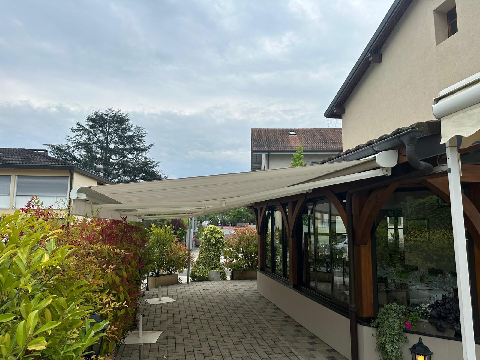 Terrasse de restaurant avec auvent beige, buissons rouges et verts et ciel nuageux.