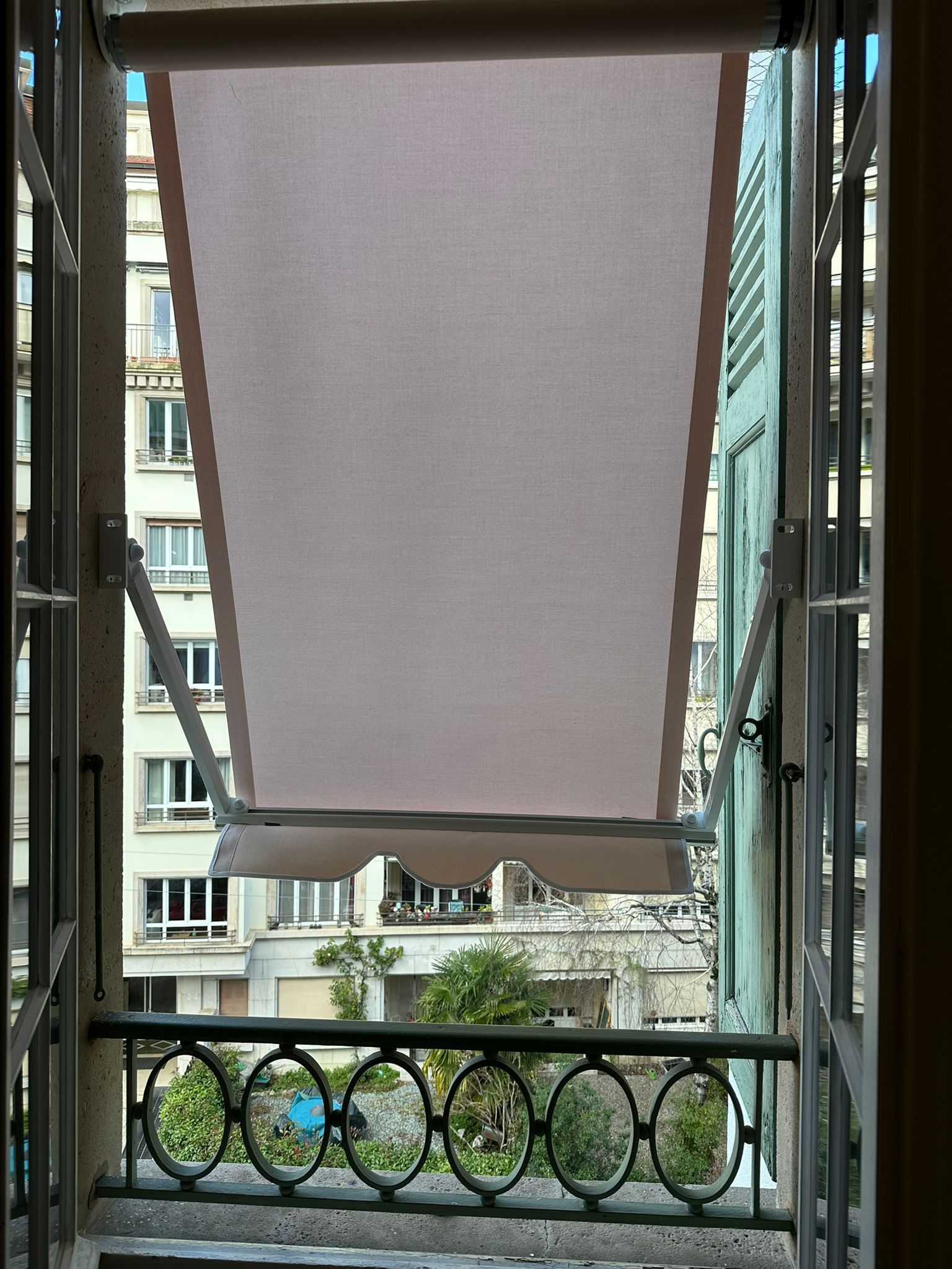 Vue d'un immeuble à travers une fenêtre avec un store beige clair partiellement tiré, avec une rampe en fer noir.