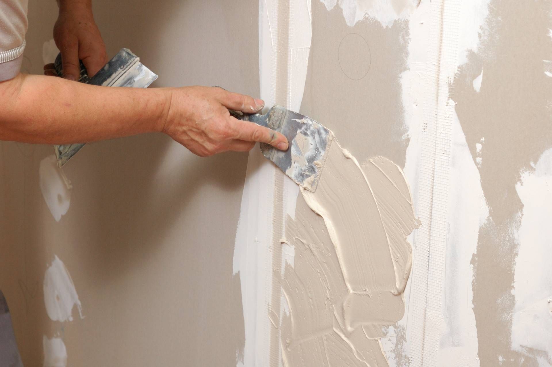 A person uses a wide metal putty knife to apply joint compound to a drywall seam on a wall.