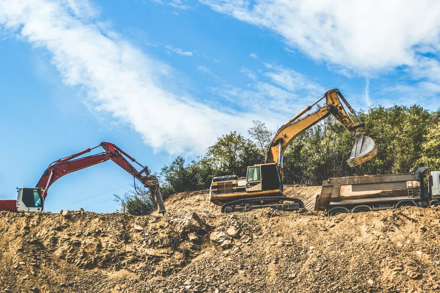 A red and a yellow excavator work on a rocky construction site, with the yellow one loading material into a truck.