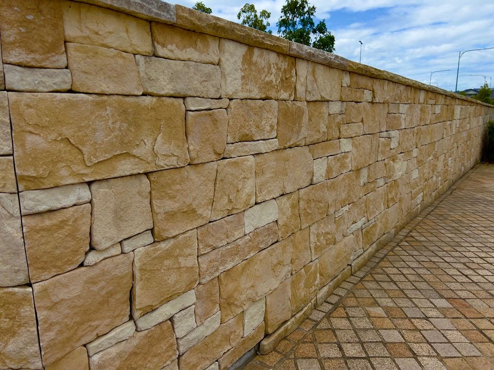 A long, waist-high wall made of rectangular, tan-colored sandstone blocks, next to a paved stone walkway.