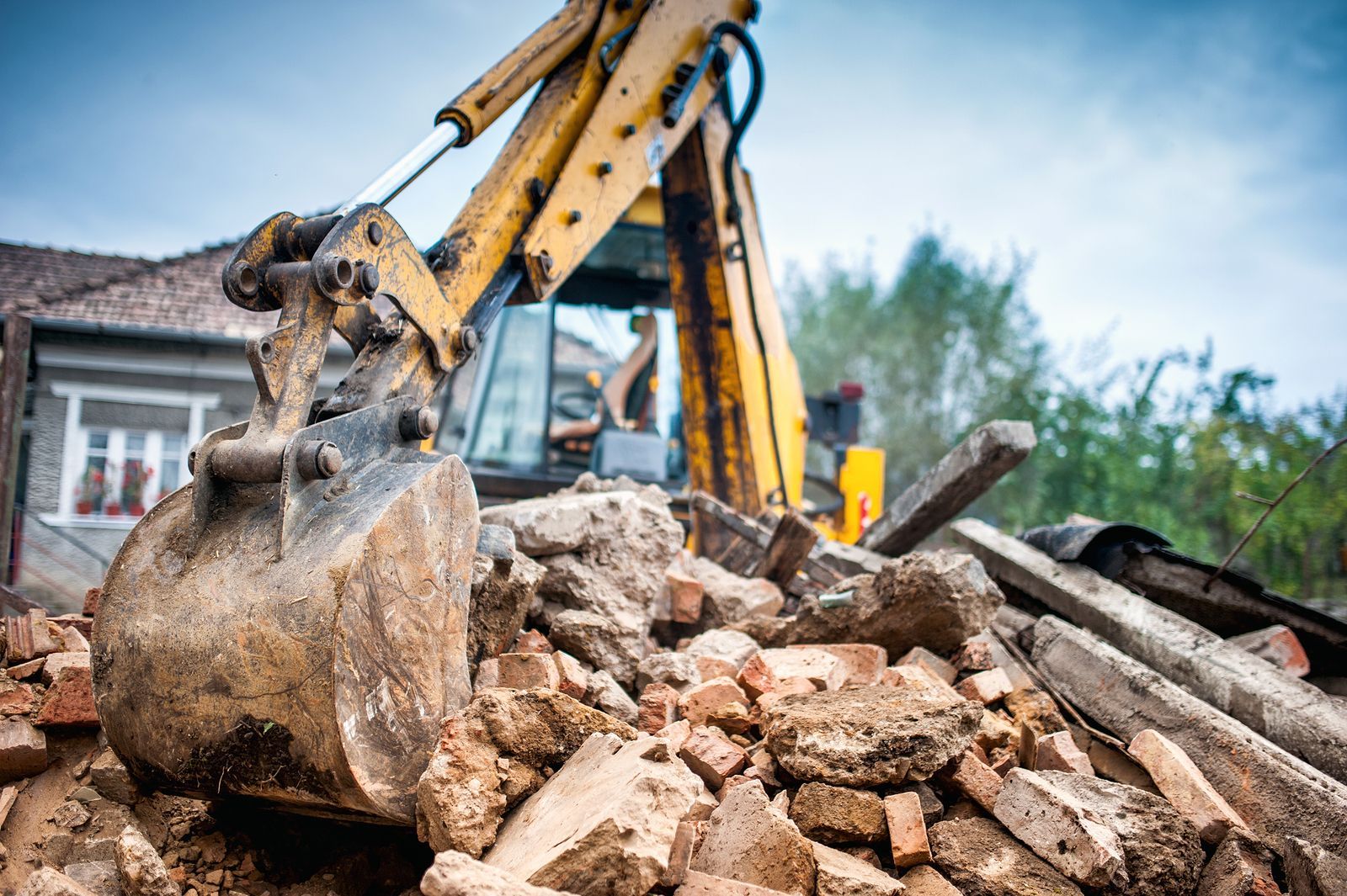 A yellow excavator bucket sits in the foreground, clearing a pile of brick and concrete rubble near a residential building.