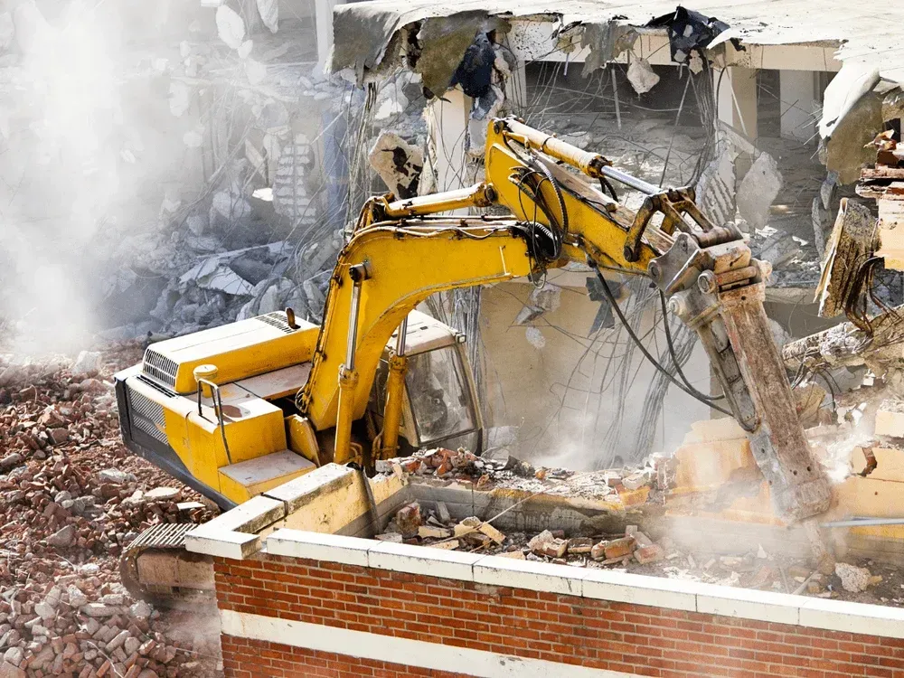 A yellow hydraulic excavator demolishes a brick building, creating a cloud of dust amidst rubble.