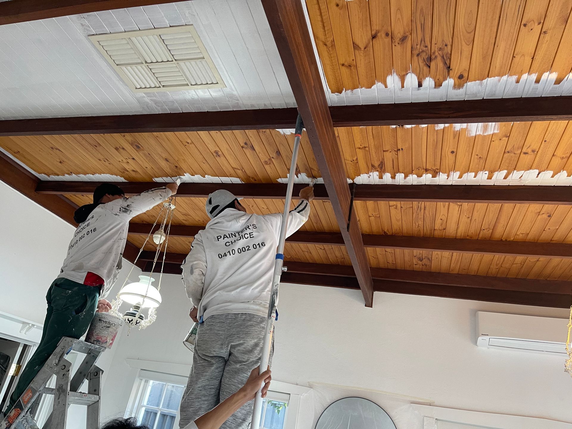 Two people are painting a wooden ceiling in a room.