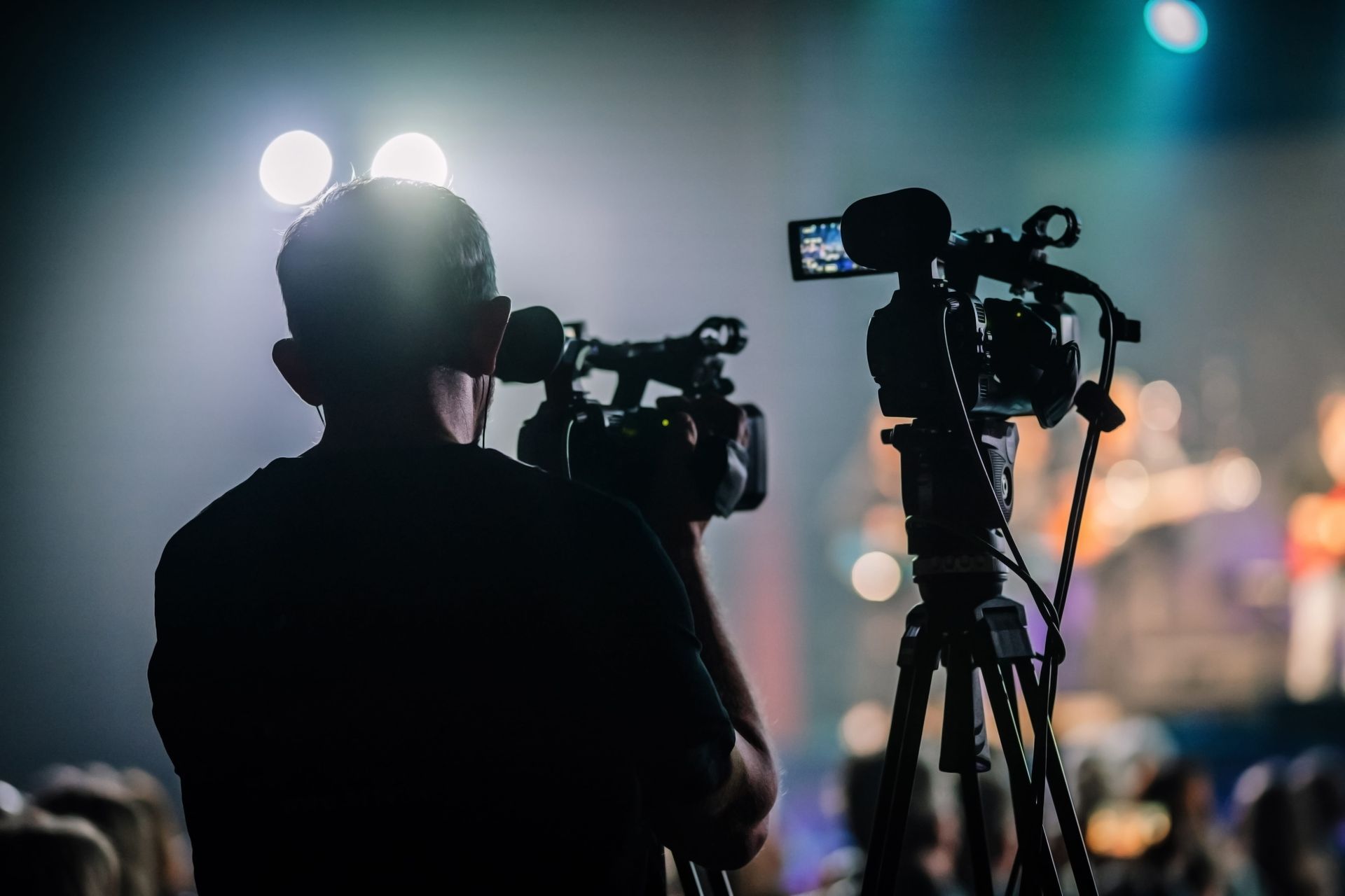Silhouetted videographer filming a live event on a tripod, with bright stage lights in the background