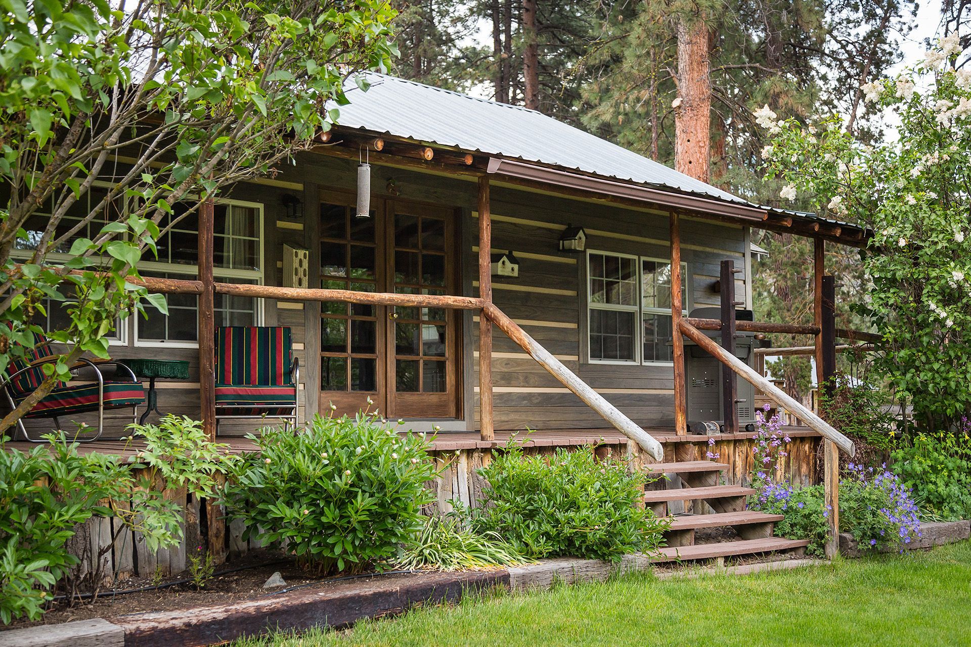 A log cabin with a porch and stairs is surrounded by trees and bushes.