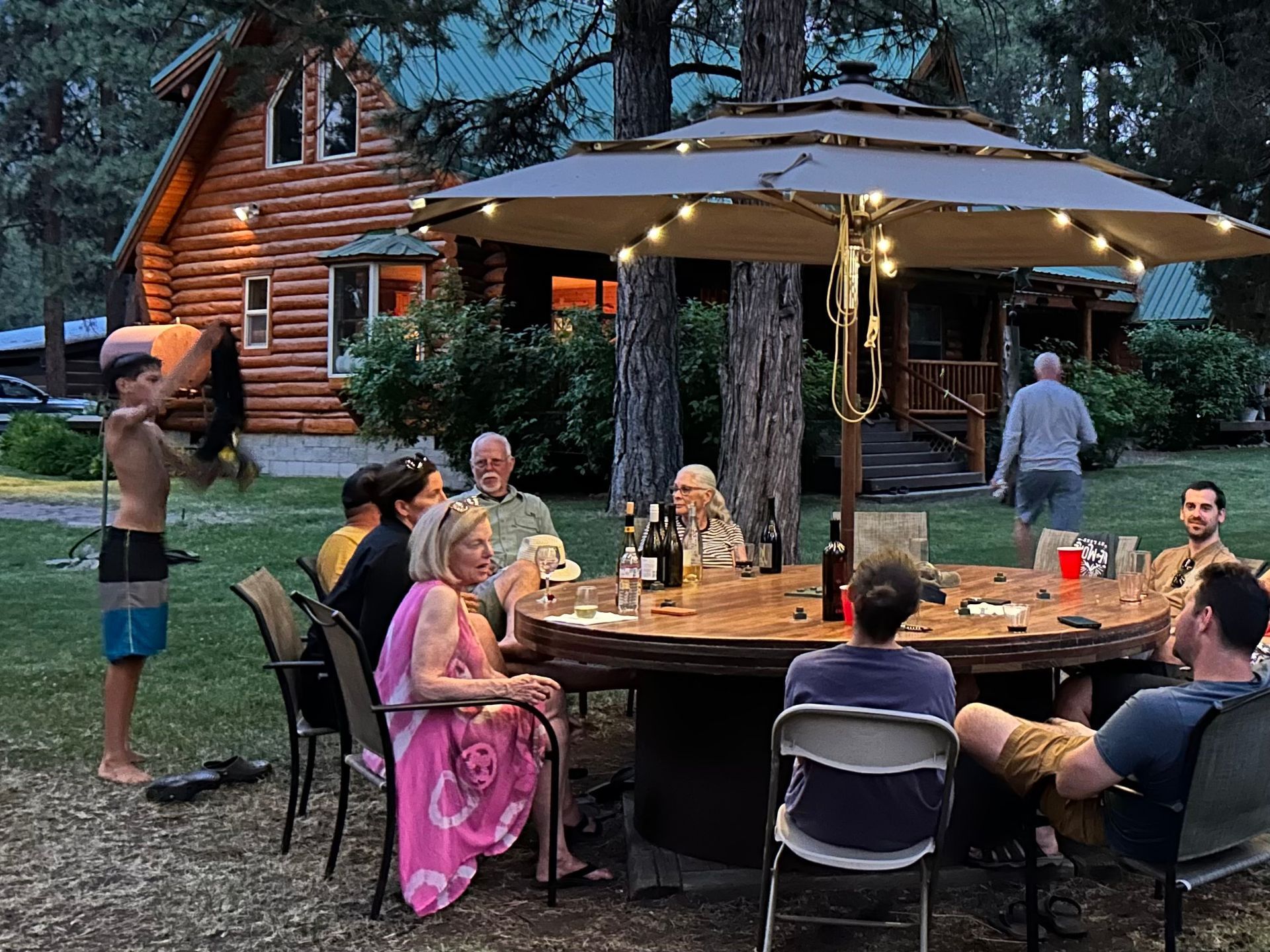 A group of people are sitting around a table under an umbrella in front of a log cabin.