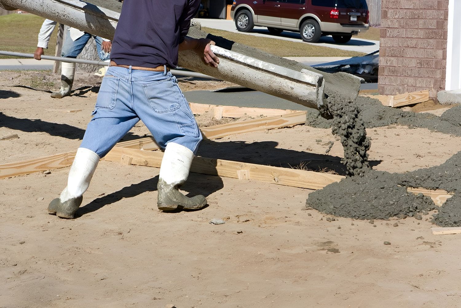 A Construction Worker In Boots Guides Wet Concrete From A Chute Onto A Sandy Work Site