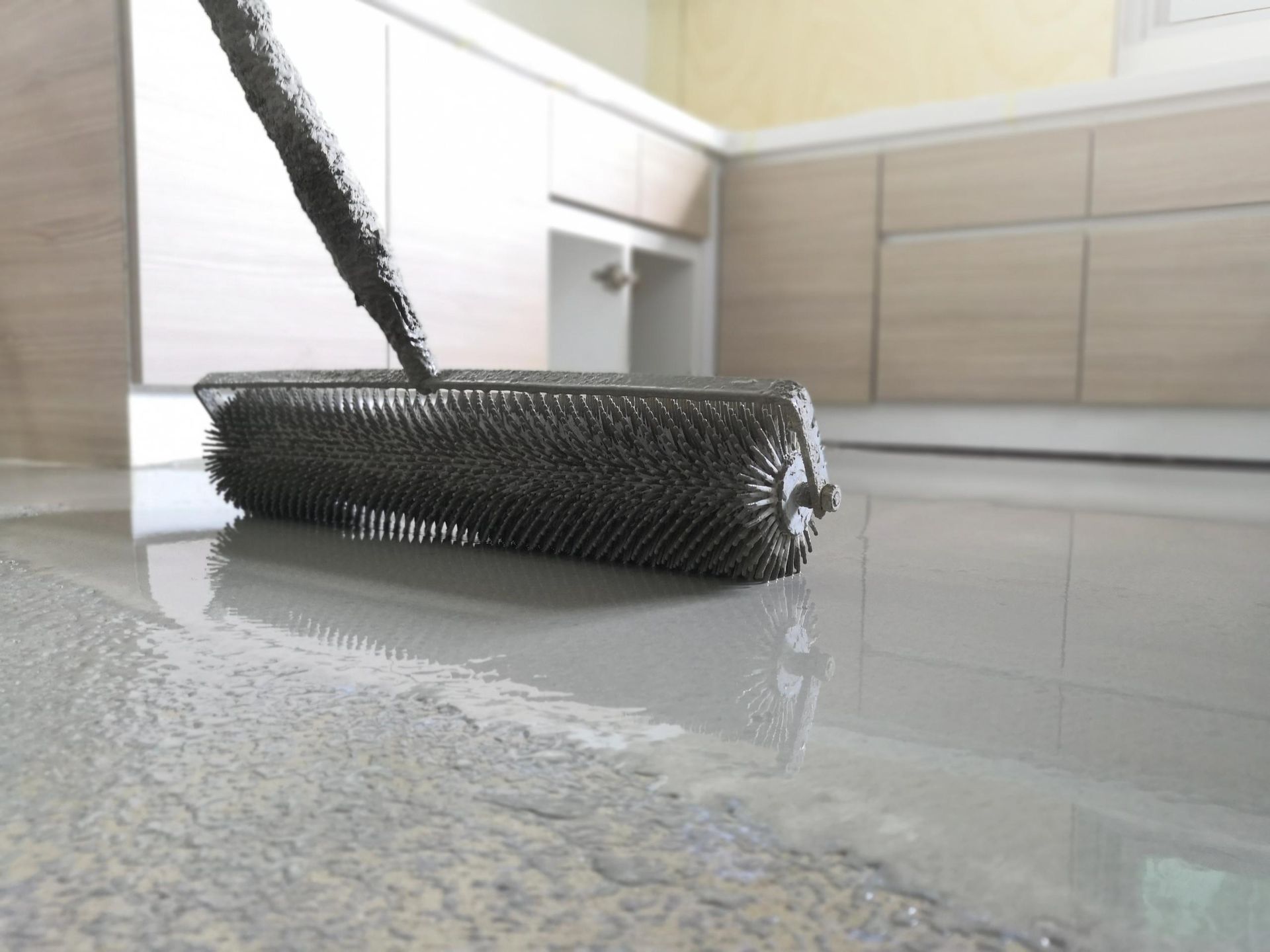 Black spiked roller tool smoothing wet, light gray epoxy floor in kitchen.