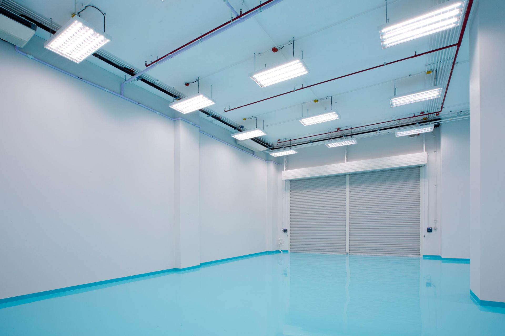 Empty, bright industrial room with white walls, a light blue floor, and a closed gray roller door.