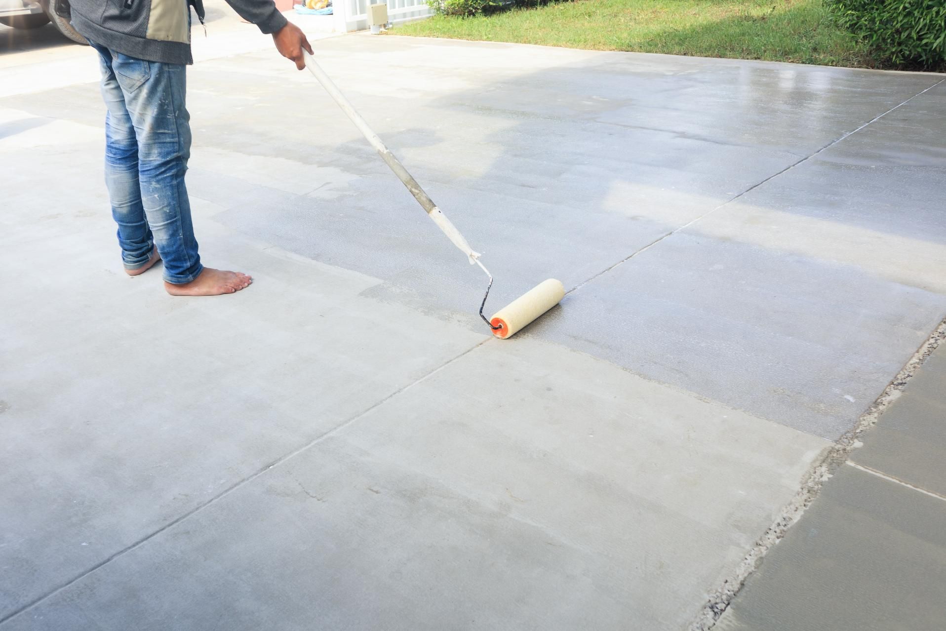 Person rolling a gray coating onto a concrete surface outdoors.