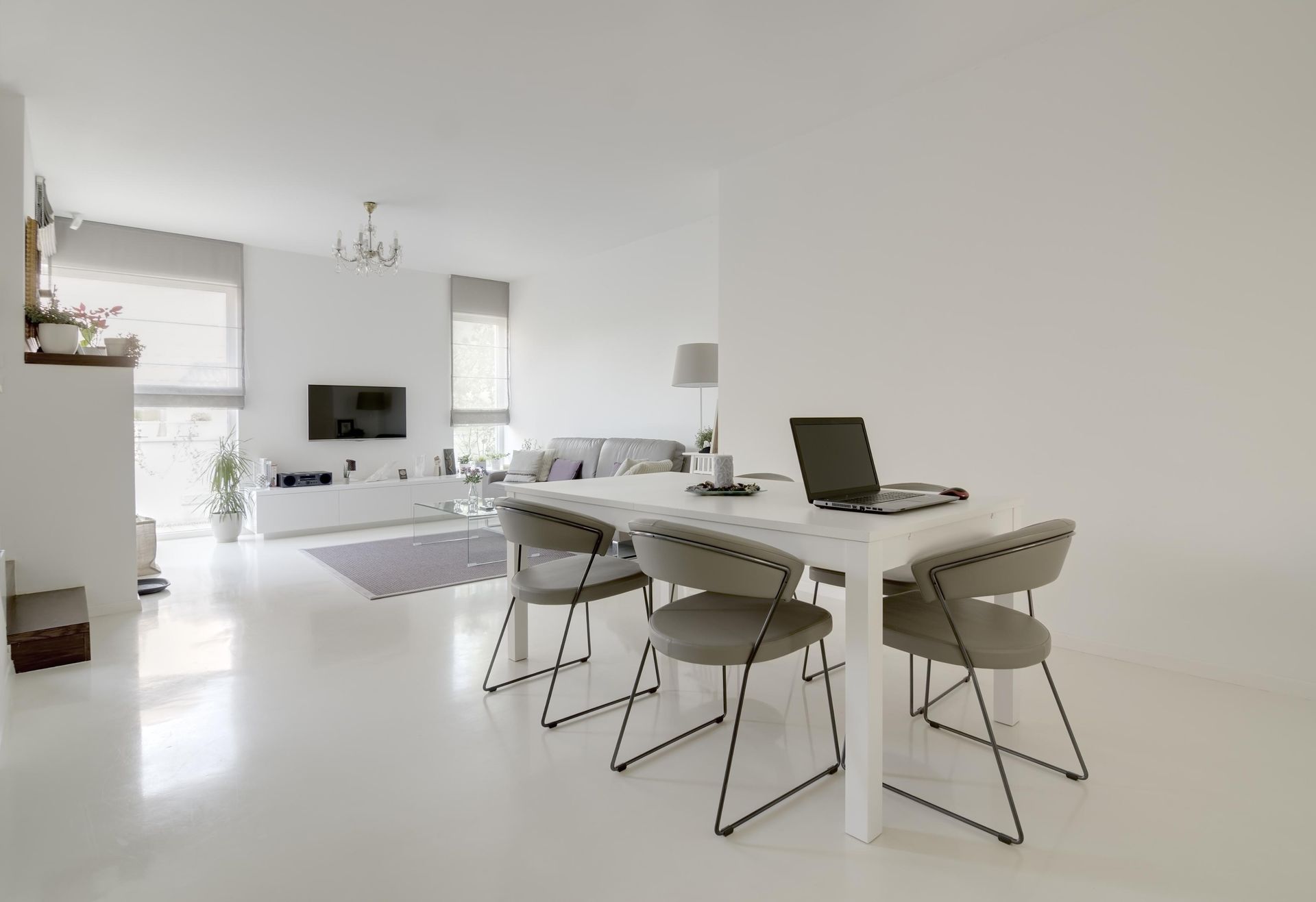 Modern, white open-concept living space with dining table, chairs, and laptop. Includes a TV, sofa, and windows.