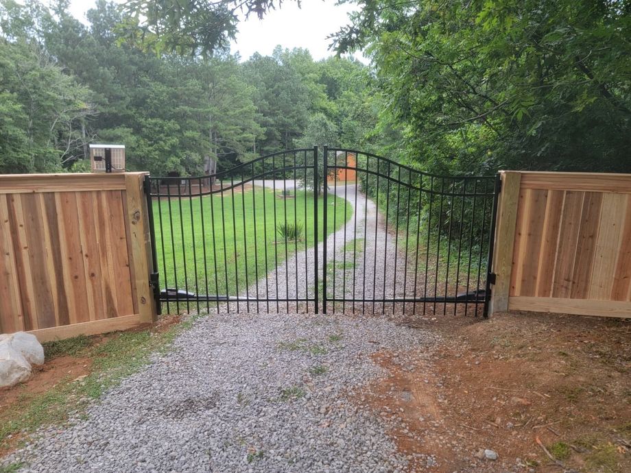 Gravel driveway leading to a closed black metal gate between wooden fences and trees.