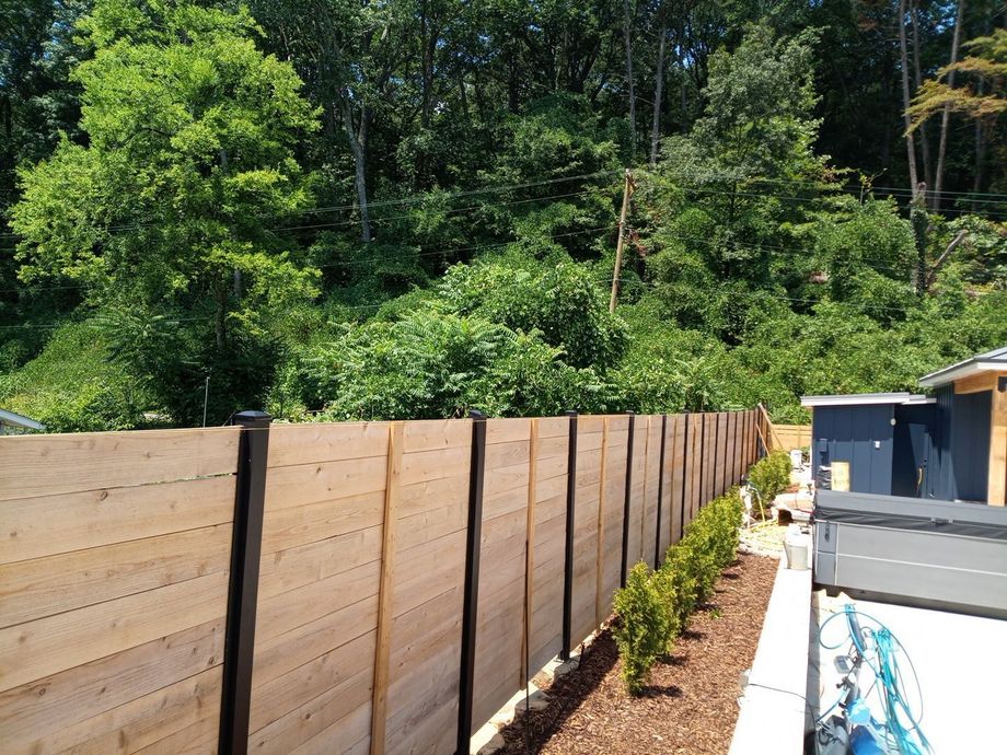 Wooden privacy fence beside a landscaped yard with young shrubs and a wooded hillside behind it