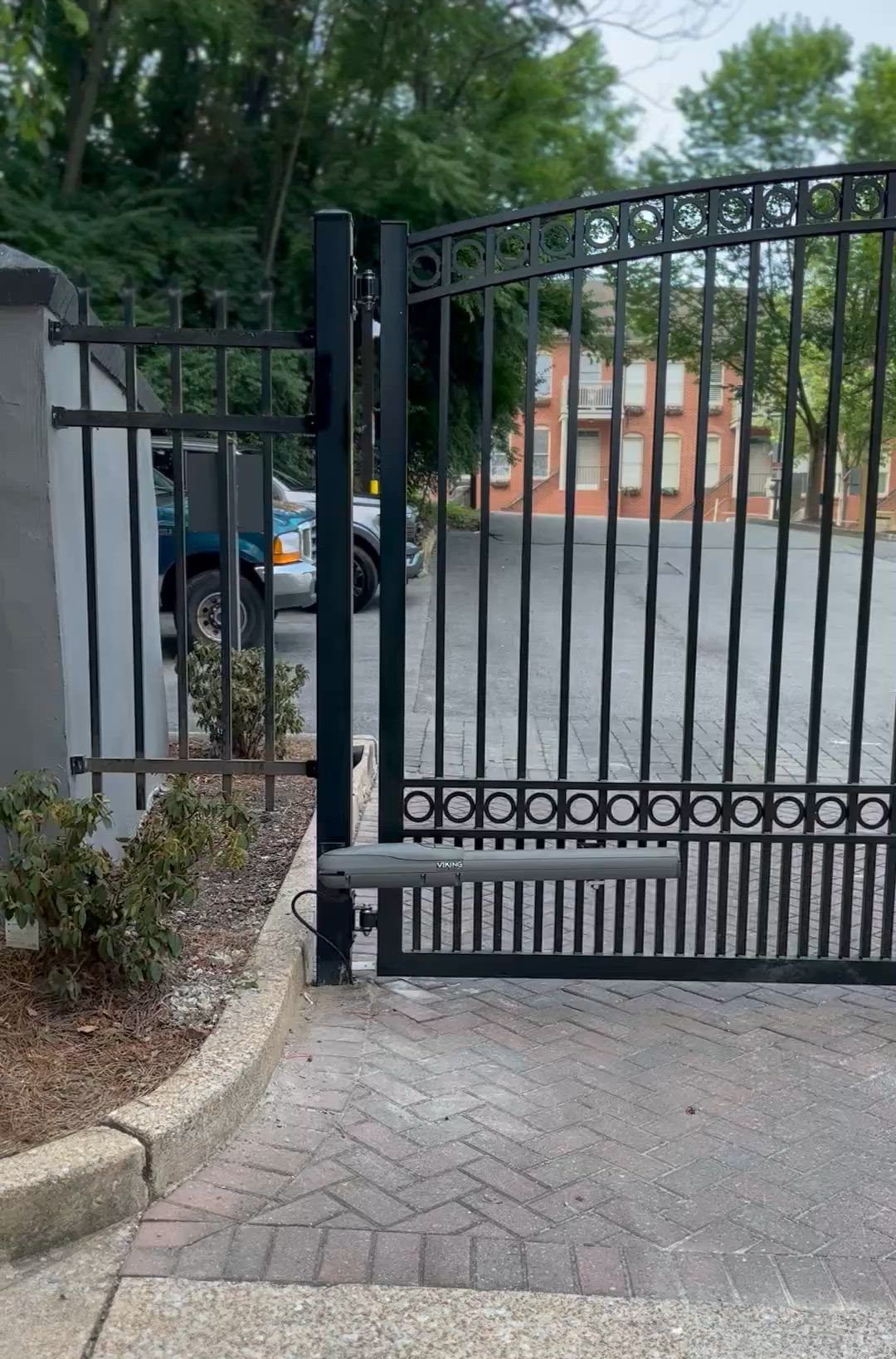 Black metal gate at a driveway entrance with trees and a parked car beyond