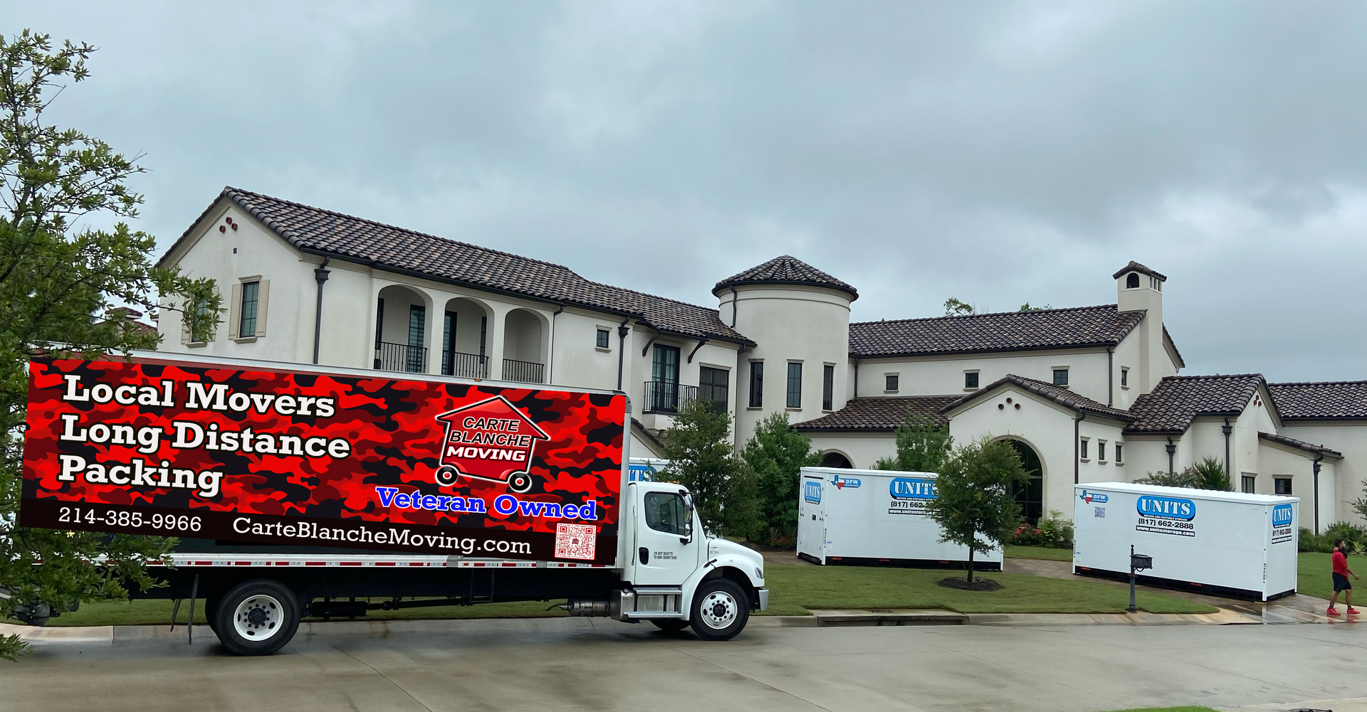 A red-branded moving truck parked in front of a large, cream-colored estate home with two smaller storage containers nearby.