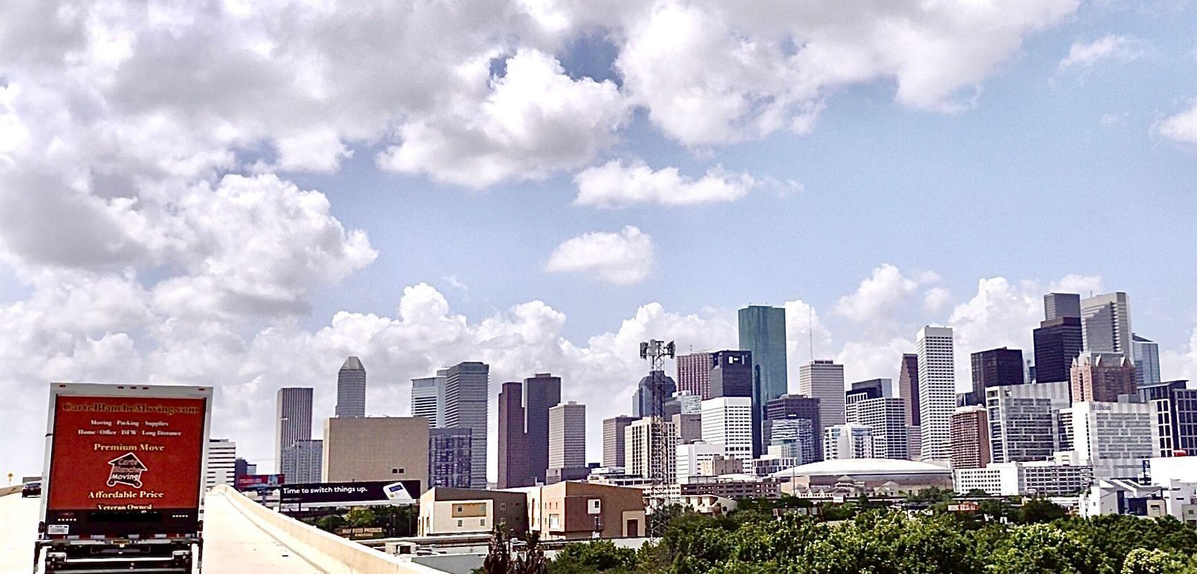 The Houston skyline under a bright, cloudy blue sky, viewed from a highway with a red truck in the foreground.