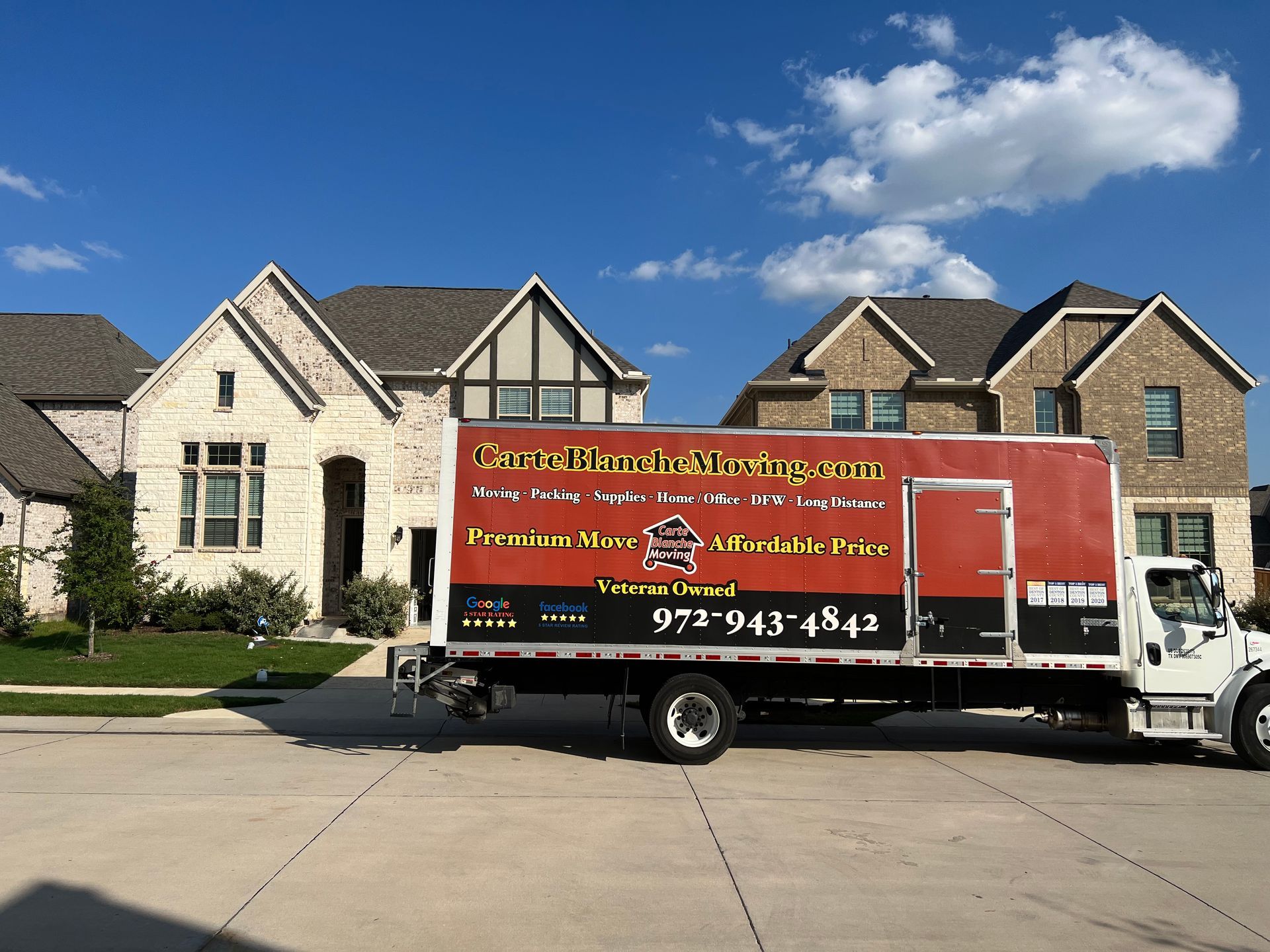 A red and black moving truck is parked in front of two large suburban houses under a blue sky with scattered clouds.