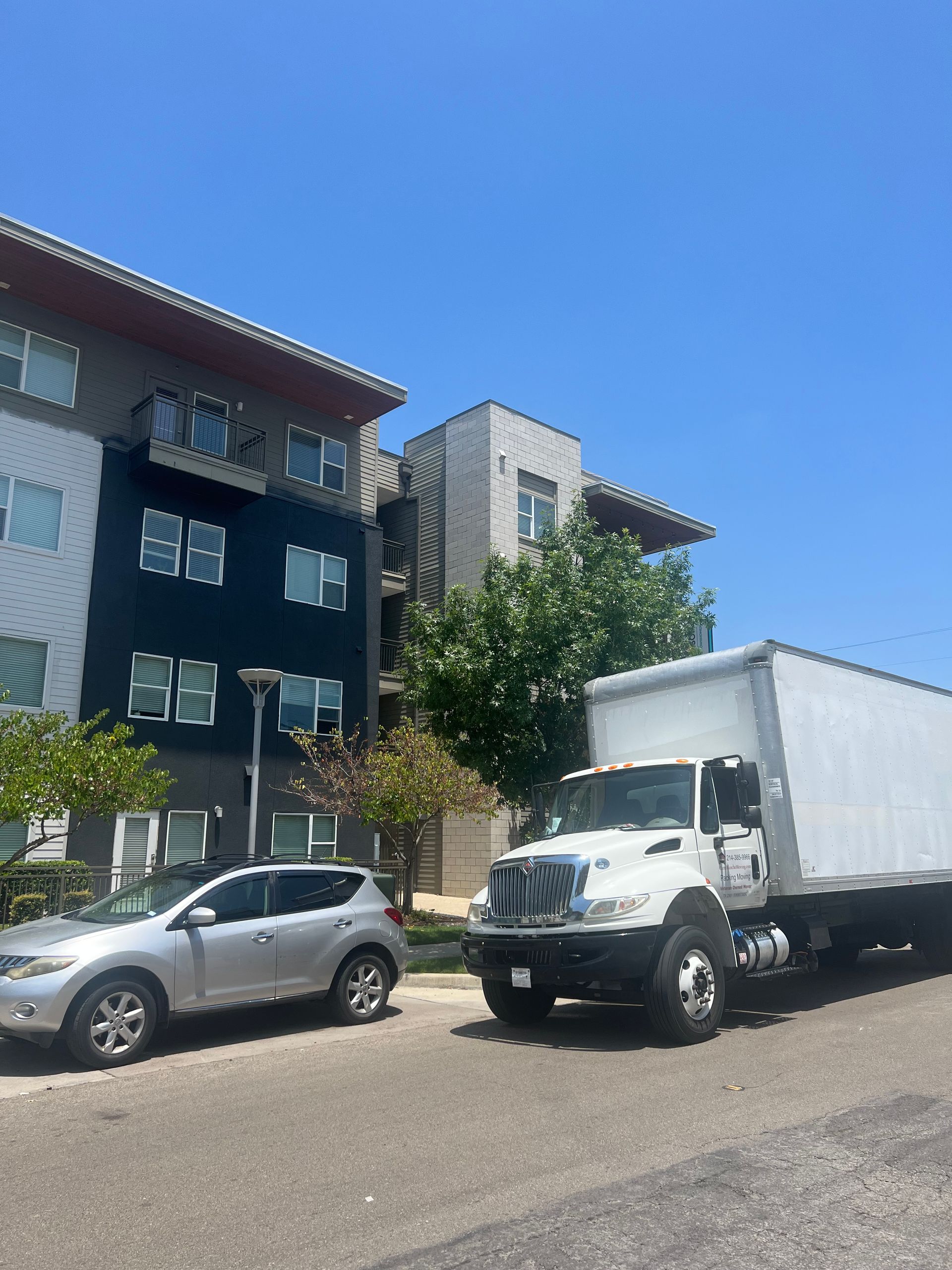 A white box truck parked on a sunny street next to a silver SUV and a modern multi-story apartment building.
